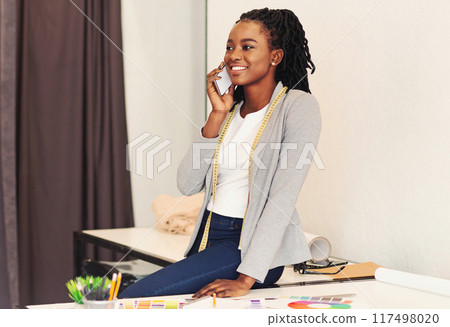 African American woman is seated at a desk, engrossed in a conversation on her cell phone. She appears focused and attentive as she communicates with the person on the other end of the line. 117498020
