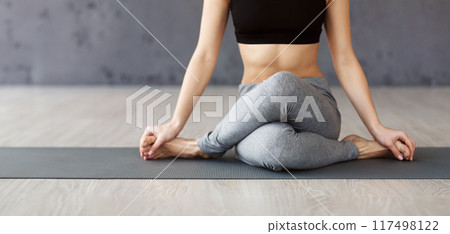 A woman is seated in a cross-legged position on a yoga mat, performing a yoga pose in a bright studio during the morning. 117498122