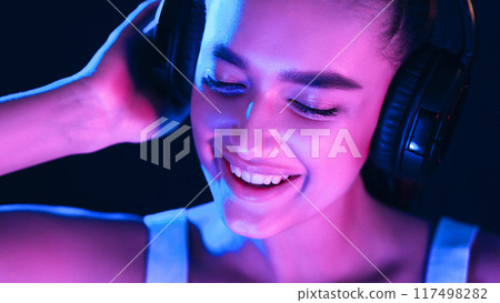 A woman is sitting while wearing headphones. She appears focused on the music she is listening to, with her eyes closed and a serene expression on her face. 117498282
