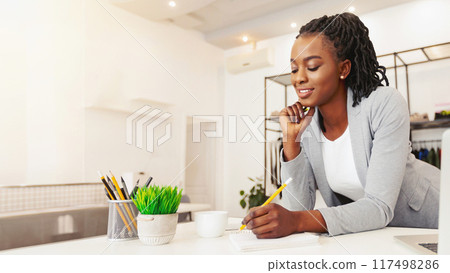 African American woman sits at a wooden desk, focusing intently as she writes on a piece of paper. Her hand holds a pen, scribbling words onto the blank sheet in front of her. African American woman sits at a wooden desk, focusing intently as she writes on a piece of paper. Her hand holds a pen, scribbling words onto the blank sheet in front of her. 117498286