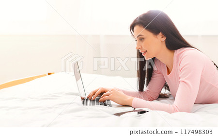 A woman is laying comfortably on a bed with a laptop computer in front of her. She appears focused and engaged in her online activities. A woman is laying comfortably on a bed with a laptop computer in front of her. She appears focused and engaged in her online activities. 117498303