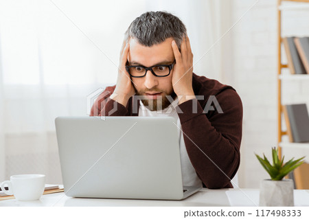 A bearded man with glasses looks shocked as he stares at a laptop screen in his home office, with bookshelves and houseplants in the background. 117498333