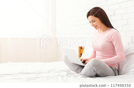 A woman is sitting on a bed, focusing on a laptop computer screen. She appears engaged in her work or leisure activity, with her hands typing on the keyboard, copy space 117498350
