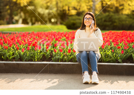 A young Asian woman with glasses is sitting on the edge of a flowerbed filled with blooming tulips while using her laptop. She appears focused and is enjoying the sunny afternoon in the park. A young Asian woman with glasses is sitting on the edge of a flowerbed filled with blooming tulips while using her laptop. She appears focused and is enjoying the sunny afternoon in the park. 117498438