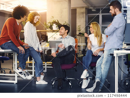 A group of four young professionals engage in a lively discussion in a brightly lit office space. One man sits on a chair holding a tablet, while the others stand around him, conversing and gesturing A group of four young professionals engage in a lively discussion in a brightly lit office space. One man sits on a chair holding a tablet, while the others stand around him, conversing and gesturing 117498444