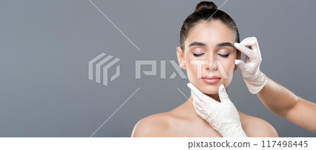 A woman is undergoing a facial lift treatment with gloves placed on her head. The gloves are used to gently lift and tighten the skin, promoting a more youthful appearance. A woman is undergoing a facial lift treatment with gloves placed on her head. The gloves are used to gently lift and tighten the skin, promoting a more youthful appearance. 117498445