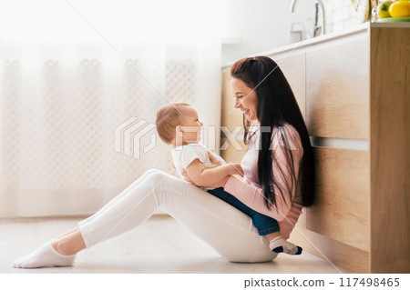A woman is seated on the floor, tenderly cradling a baby in her arms. Both individuals are looking at each other, creating a touching moment of connection and care. 117498465