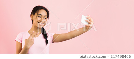 African American girl is standing in front of a pink background, holding a smartphone and posing for a selfie. She looks into the camera with a smile as she captures the moment. African American girl is standing in front of a pink background, holding a smartphone and posing for a selfie. She looks into the camera with a smile as she captures the moment. 117498466