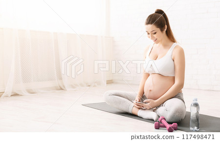 A pregnant woman is seated on a yoga mat, engrossed in her phone. She appears focused as she scrolls through the screen. Her bump is prominent, indicating her stage of pregnancy. A pregnant woman is seated on a yoga mat, engrossed in her phone. She appears focused as she scrolls through the screen. Her bump is prominent, indicating her stage of pregnancy. 117498471