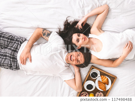 A happy couple lies in bed together, enjoying a breakfast tray with coffee, croissants, and fruit. The woman is looking towards the camera, while the man smiles, looking at his partner. A happy couple lies in bed together, enjoying a breakfast tray with coffee, croissants, and fruit. The woman is looking towards the camera, while the man smiles, looking at his partner. 117498488