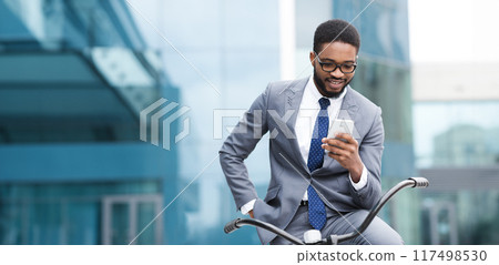 A professional black man is outdoors with his bike, using his phone. The scene is calm and urban, reflecting a balance of professional duties and leisure activities. 117498530