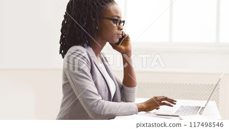 Concentrated black lady CEO multitasking at modern office interior, checking data on her laptop while chatting on phone, copy space, side view Concentrated black lady CEO multitasking at modern office interior, checking data on her laptop while chatting on phone, copy space, side view 117498546