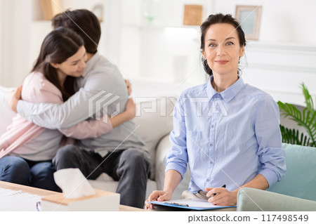 A couple hugs each other during a morning counseling session while a therapist takes notes and offers a confident smile in a bright, well-lit room. 117498549