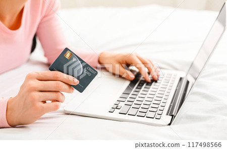 Cropped of lady laying on a bed while using a laptop and holding a credit card, clearly engaging in online shopping or digital payment Cropped of lady laying on a bed while using a laptop and holding a credit card, clearly engaging in online shopping or digital payment 117498566
