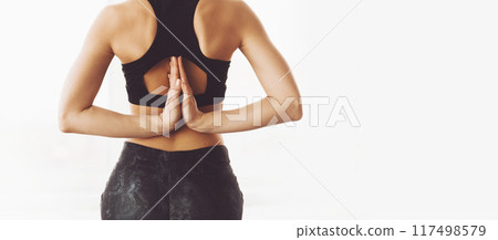 A woman stands against a stark white backdrop, her body garbed in dark, form-fitting yoga attire. Her hands are pressed together in a traditional Namaste gesture, positioned at her back 117498579
