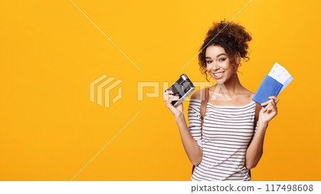 A cheerful young African American woman with curly hair is standing against a bright orange backdrop, holding up her passport and a camera, suggesting excitement about an upcoming travel, copy space 117498608