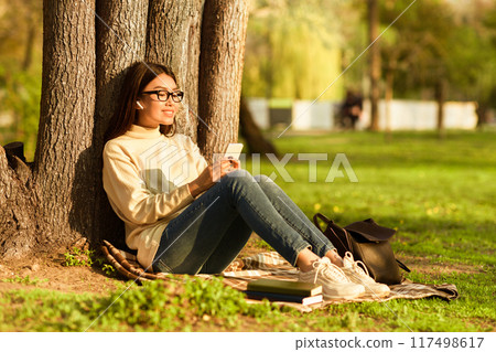 Asian young woman is relaxing under a tree in a sunny park, looking at her smartphone. She is wearing casual clothes and glasses, sitting comfortably on a blanket with books beside her 117498617