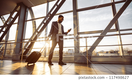 A professional black man with luggage is on his phone at the airport. The atmosphere is dynamic and modern, reflecting business travel and connectivity, sun flare A professional black man with luggage is on his phone at the airport. The atmosphere is dynamic and modern, reflecting business travel and connectivity, sun flare 117498627