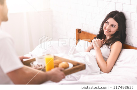 A man is bringing a tray of breakfast in bed to a woman who is sitting up in bed and smiling. The tray contains a glass of orange juice, a cup of coffee, and some pastries. A man is bringing a tray of breakfast in bed to a woman who is sitting up in bed and smiling. The tray contains a glass of orange juice, a cup of coffee, and some pastries. 117498635