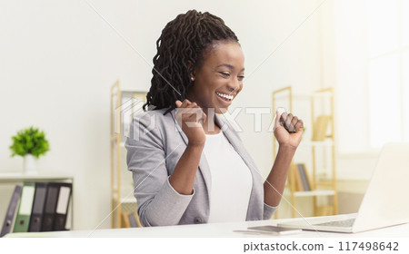 A joyful African American woman employee is working on a computer at her desk. The office is modern and tidy, capturing her happiness and excitement as she focuses on her tasks. 117498642