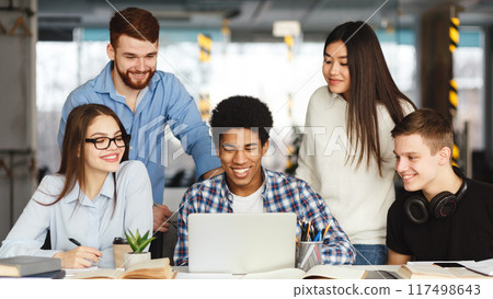 A diverse group of people are gathered around a table indoors. They are actively engaged and focused on a laptop screen, likely discussing work-related matters or collaborating on a project. 117498643
