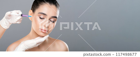 A doctor wearing white gloves is administering a cosmetic injection to a young woman face. The woman appears calm and content as she closes her eyes during the procedure, copy space A doctor wearing white gloves is administering a cosmetic injection to a young woman face. The woman appears calm and content as she closes her eyes during the procedure, copy space 117498677
