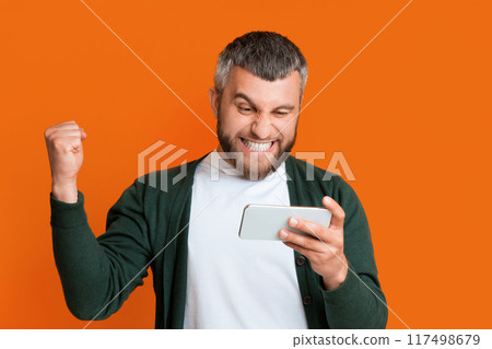 A man in casual clothing holding a smartphone in one hand and raising his fist in a celebratory gesture. The man appears focused and confident. A man in casual clothing holding a smartphone in one hand and raising his fist in a celebratory gesture. The man appears focused and confident. 117498679