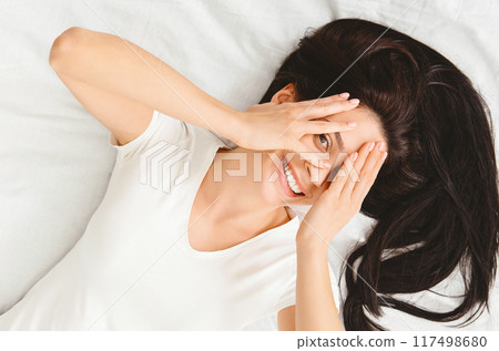 Playful young woman hiding face, feeling shy, peeking through fingers, lying cozy in bed, top view 117498680