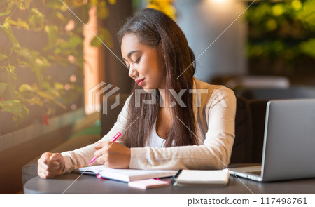 A young black girl is seated at a table, concentrating as she writes in a notebook. Her pen moves swiftly across the paper, capturing her thoughts and ideas in the quiet setting. A young black girl is seated at a table, concentrating as she writes in a notebook. Her pen moves swiftly across the paper, capturing her thoughts and ideas in the quiet setting. 117498761