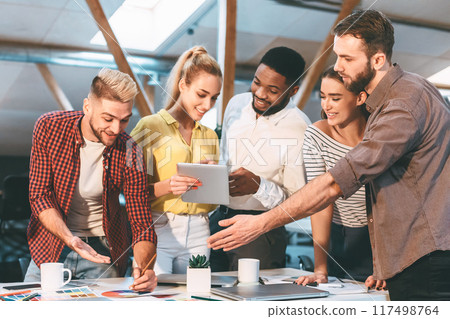 Five colleagues gather around a table in an office, discussing and collaborating on a project using documents and a tablet. Five colleagues gather around a table in an office, discussing and collaborating on a project using documents and a tablet. 117498764