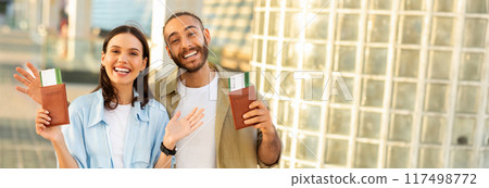 Happy young european guy with beard and woman hold passports and tickets, enjoy trip to station, outdoor. Tourist emotion, active lifestyle, travel at summer, vacation together, copy space Happy young european guy with beard and woman hold passports and tickets, enjoy trip to station, outdoor. Tourist emotion, active lifestyle, travel at summer, vacation together, copy space 117498772