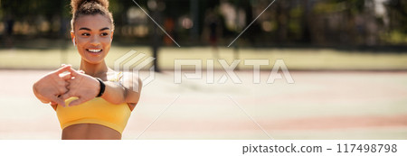 Portrait of sporty African American woman in wireless earbuds warming up before jogging at park, stretching her arms muscles outdoors, feeling healthy and motivated, banner, panorama, free copy space 117498798