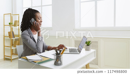 Elated young African American woman professional in formal wear, working on a laptop in the office, talking on the phone. Elated young African American woman professional in formal wear, working on a laptop in the office, talking on the phone. 117498800