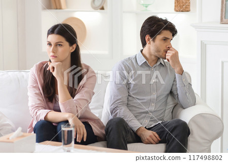 A young couple sits on a sofa in a brightly lit living room. Both appear to be lost in thought, staring off into the distance with serious expressions. Crisis in relationships A young couple sits on a sofa in a brightly lit living room. Both appear to be lost in thought, staring off into the distance with serious expressions. Crisis in relationships 117498802