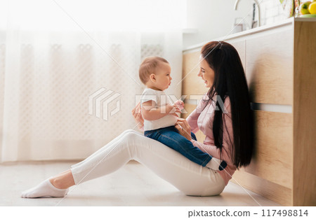 A woman is seated on the floor, cradling a baby in her arms. She gazes down at the baby with a tender expression, providing comfort and care in a cozy setting. A woman is seated on the floor, cradling a baby in her arms. She gazes down at the baby with a tender expression, providing comfort and care in a cozy setting. 117498814