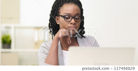 African American woman is focused on her screen in a modern office. The scene is professional and sleek, with a tidy desk and bright lighting, emphasizing her concentration. 117498829