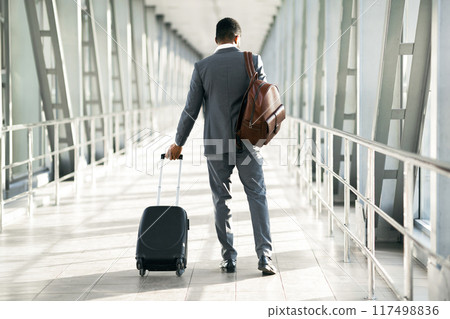 A corporate traveler with luggage is seen at an airport. The mood is focused and professional, with a modern airport setting that emphasizes travel and business, back view A corporate traveler with luggage is seen at an airport. The mood is focused and professional, with a modern airport setting that emphasizes travel and business, back view 117498836