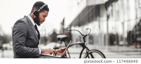 African American businessman using headphones and laptop outdoors, sitting on concrete bench at downtown, bicycle by him, side view, copy space African American businessman using headphones and laptop outdoors, sitting on concrete bench at downtown, bicycle by him, side view, copy space 117498849