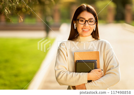 Asian young woman wearing glasses and earbuds is seen walking on a college campus, holding several books and smiling warmly during a sunny day. 117498858