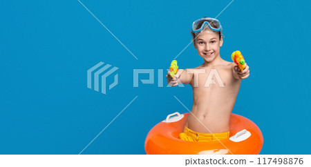 A young boy stands against a blue background, wearing swimming goggles and a colorful swim ring, enthusiastically aiming two water guns forward, copy space A young boy stands against a blue background, wearing swimming goggles and a colorful swim ring, enthusiastically aiming two water guns forward, copy space 117498876
