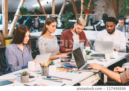 A group of young professionals work together in a modern, open-office environment with natural light, laptops, and design materials. A group of young professionals work together in a modern, open-office environment with natural light, laptops, and design materials. 117498886