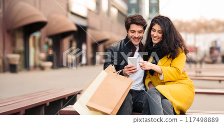 A man and woman are seated on a bench, holding multiple shopping bags, surrounded by a cityscape. 117498889