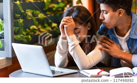 A concerned black guy teenager offers comfort to a distressed friend who is sitting with her head in her hands at a cafe. Open notebooks and a laptop are on the table A concerned black guy teenager offers comfort to a distressed friend who is sitting with her head in her hands at a cafe. Open notebooks and a laptop are on the table 117498892