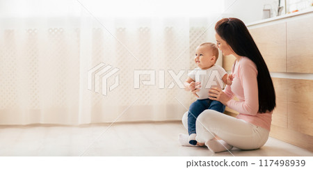A woman is seated on the floor next to a baby, both engaged in a moment of connection and interaction. The woman appears to be gently tending to the baby, who is calmly looking at her, copy space 117498939