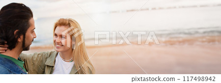 A man and woman are standing close together on a beach at sunset. The man has dark hair and is wearing a blue shirt. The woman has blonde hair and is wearing a green jacket, copy space A man and woman are standing close together on a beach at sunset. The man has dark hair and is wearing a blue shirt. The woman has blonde hair and is wearing a green jacket, copy space 117498942