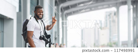 A young African American man with a beard and a black backpack smiles and gives a peace sign while holding a camera, standing in an urban environment with a building in the background, copy space 117498991