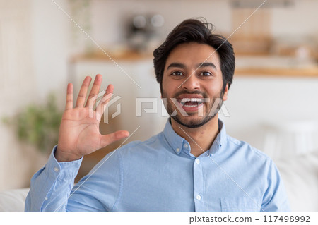 A young man with dark hair and a beard is wearing a blue button-down shirt and smiling broadly. He is waving with his right hand, extending all five fingers toward the camera. 117498992