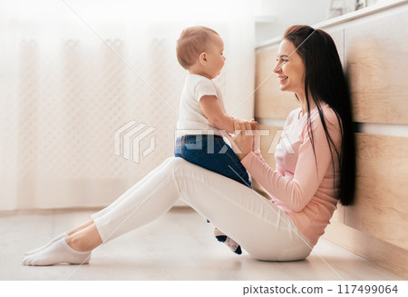 A woman is seated on the floor, cradling a baby in her arms. She gazes down at the infant with a gentle expression, providing care and comfort in a nurturing embrace. A woman is seated on the floor, cradling a baby in her arms. She gazes down at the infant with a gentle expression, providing care and comfort in a nurturing embrace. 117499064