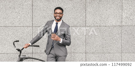 Black guy professional is riding his bicycle to work, holding a cup of takeaway coffee. The atmosphere is urban and energetic, showing a blend of efficiency and casual commuting. 117499092