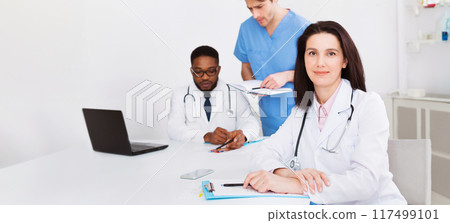 Female doctor at concilium, looking at camera. Medical team on background Female doctor at concilium, looking at camera. Medical team on background 117499101